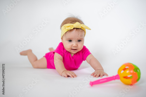 cute, beautiful little girl in a pink bodysuit and a bandage lies on a white background, smiles and gnaws toys. beautiful baby. teeth are erupting. portrait of a baby. portrait of a beautiful girl