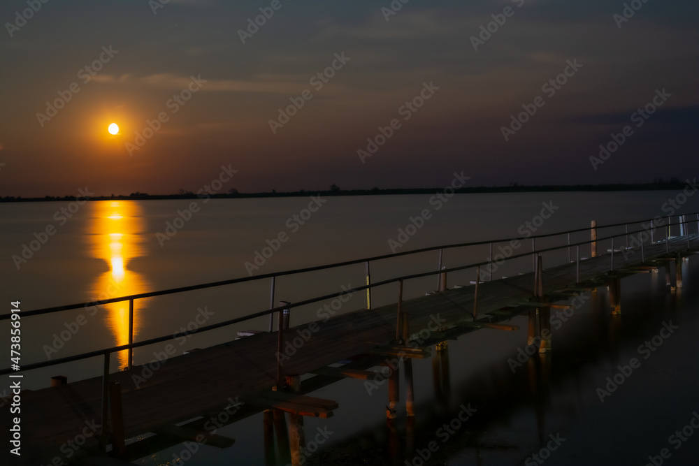 A wooden pedestrian bridge with railings runs along the lake, around the shallow water. There are bright stars and the moon in the sky. A wooden pedestrian bridge with railings runs along the lake, around the shallow water. There are bright stars and the moon in the sky.