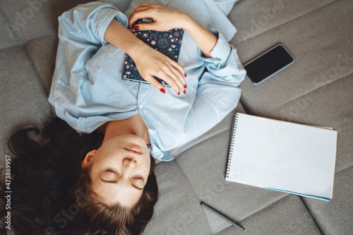 A young woman is lying at home on the sofa with her eyes closed, a diary in her hands and dreaming.