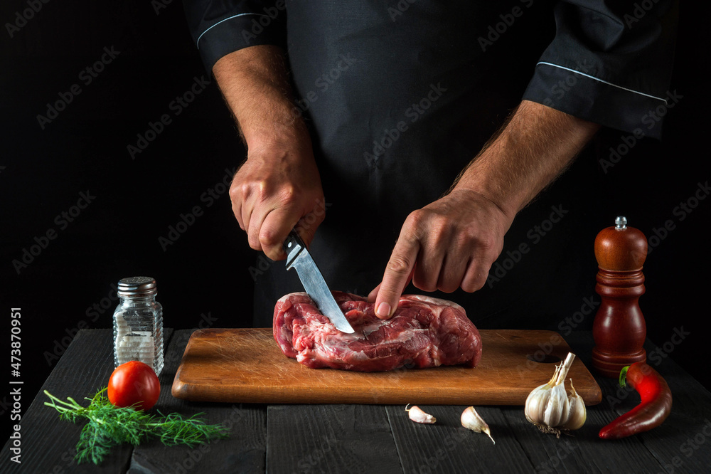 Cook cuts meat with a knife in the kitchen prepares food. Vegetables and spices on the kitchen table in a restaurant to prepare a delicious lunch