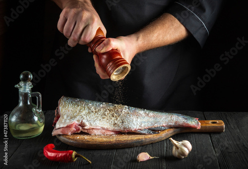 Professional chef prepares fresh fish sprinkling pepper. Preparing to cook fish food. Working environment in restaurant or cafe kitchen photo