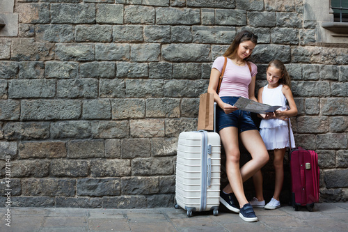 Young cheerful positive woman and little girl traveler with suitcase leaning against stone wall