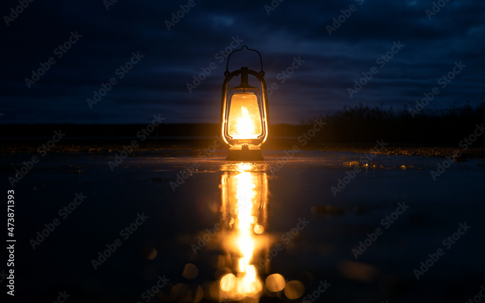 Beautiful colorful illuminated lantern on frozen puddle. The lamp is beautifully reflected in the mirror surface. Cloudy sky.