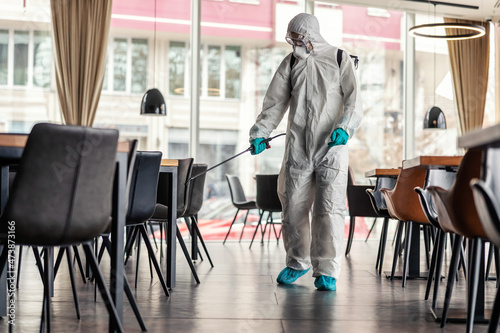 COVID 19 warning. A man in protective white clothing and a sprayer prevents the spread of the infection in a restaurant place. Stay responsible, COVID 19 alert © dusanpetkovic1