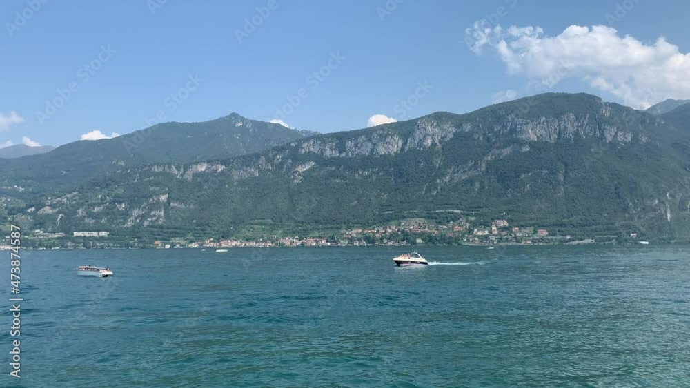 Speed boat slowly goes, sails in blue Lago di Como Lake. Varenna town and the mountains in the background. It is a famous sightseeing and tourism place. Bellagio, Como Lake, Lombardy, Italy Speed boat slowly goes, sails in blue Lago di Como Lake. Varenna town and the mountains in the background. It is a famous sightseeing and tourism place. Bellagio, Como Lake, Lombardy, Italy