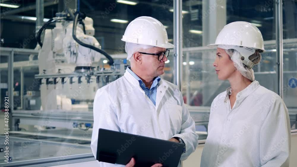 Two engineers in hardhats are talking next to robotic machines Two engineers in hardhats are talking next to robotic machines