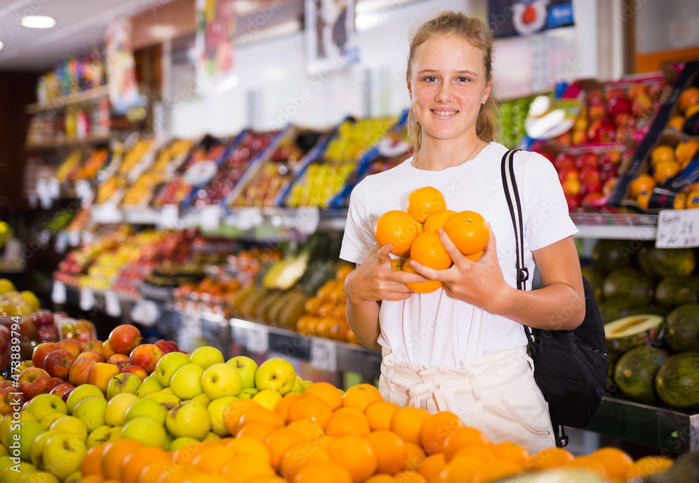 Portrait of a smiling fifteen-year-old girl in a store, standing near a fruit counter, holding oranges in her hands
