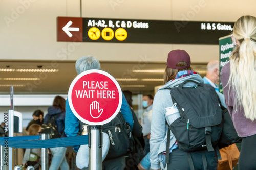 Travelers wait in line at a busy airport during covid next to a please wait here sign.