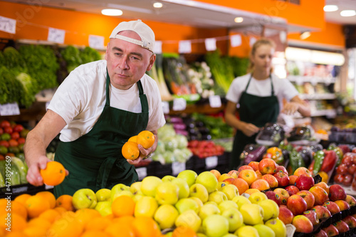 Portrait of cheerful male seller in uniform holding fresh oranges in grocery shop