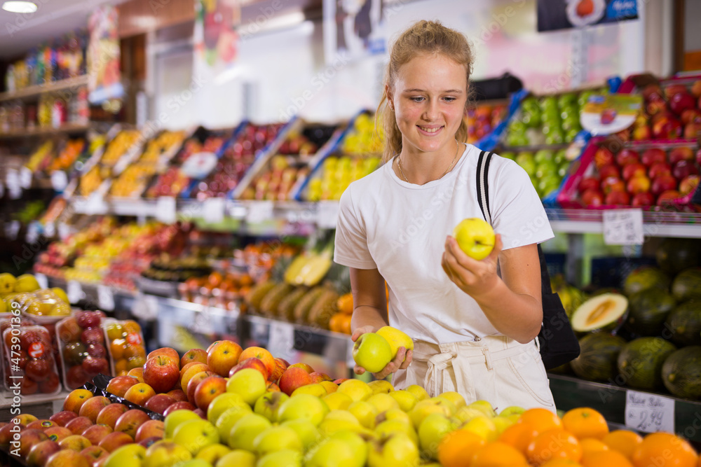 Positive girl consumer choosing fresh apples at grocery section of supermarket