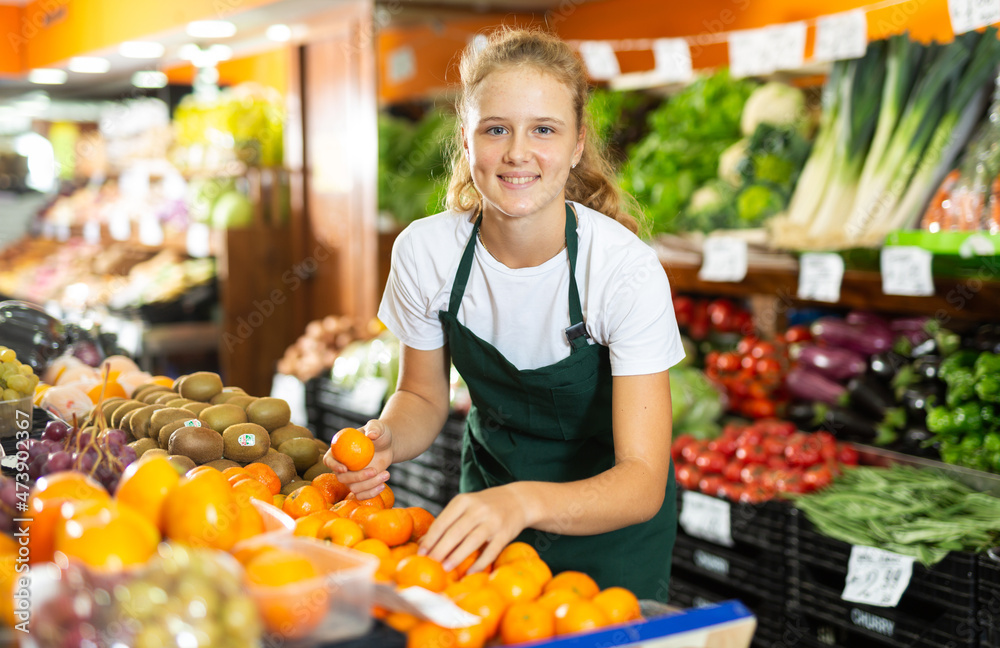 Obraz premium Fifteen-year-old girl who works part-time in a store as a trainee saleswoman puts tangerines on the counter for sale