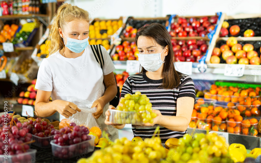 Young woman with a fifteen-year-old girl in protective masks, who came to the supermarket for shopping during the pandemic, ..choose grapes at the counter