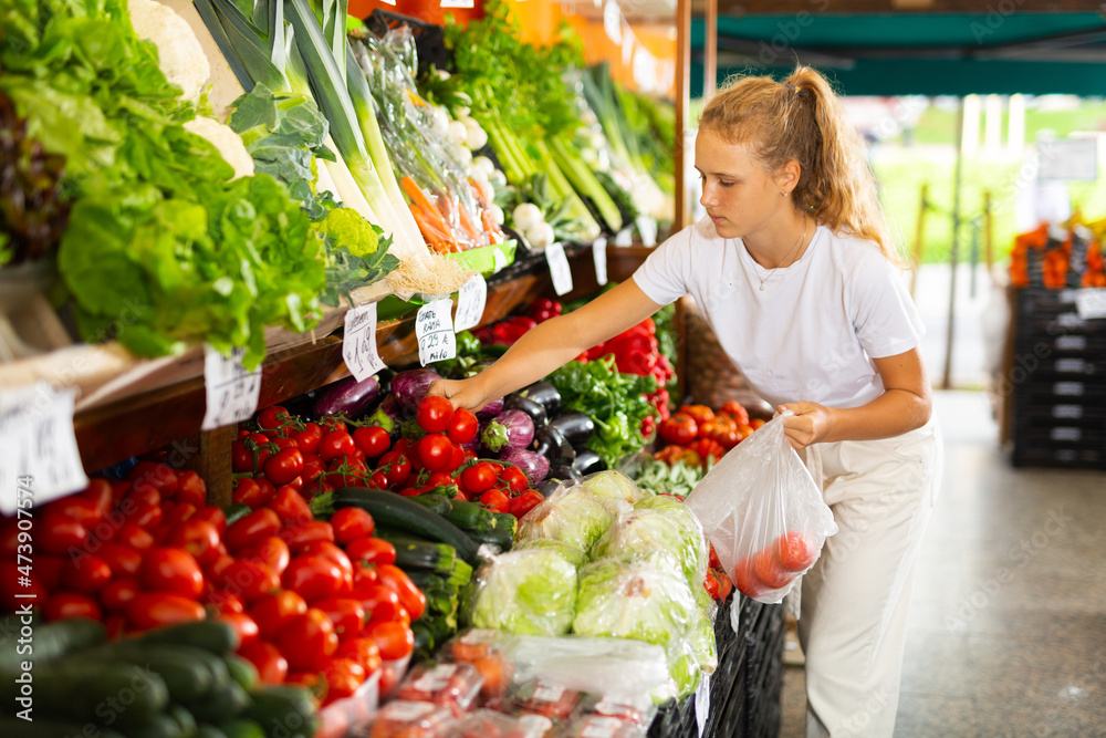 Positive teenage girl choosing fresh tomato at grocery section of supermarket