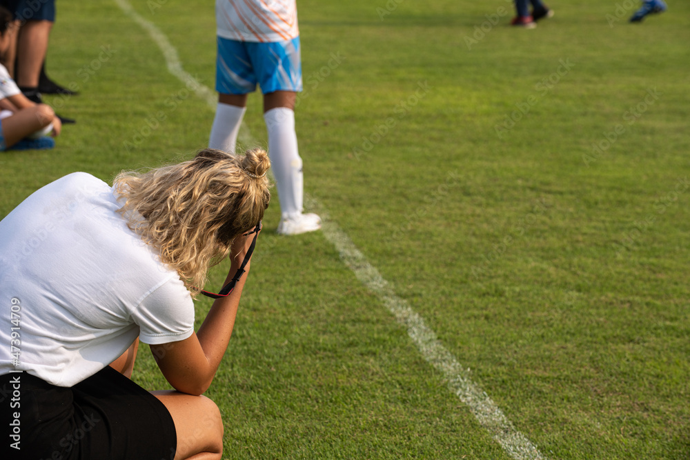 Mother taking pictures of her daughter playing football in a high school tournament on a clear sky and sunny day. Sport, active lifestyle, happy family and soccer mom concept.