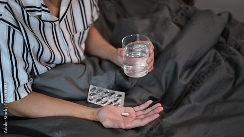 Cropped shot young man holding pill and glass of water while sitting on bed.