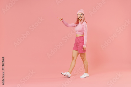 Full body smiling fun young woman 20s with bright dyed rose hair in rosy top shirt hat walking going point index finger aside on workspace area isolated on plain light pastel pink background studio.