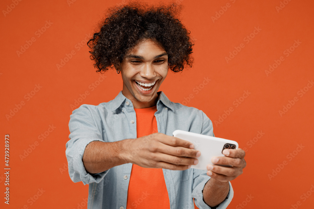 Young gambling black man 50s wear blue shirt t-shirt using play racing app on mobile cell phone hold gadget smartphone for pc video games isolated on plain orange background. People lifestyle concept.