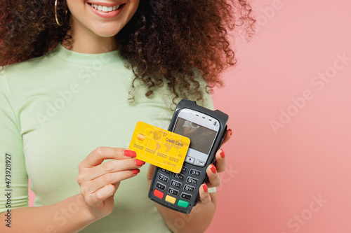 Close up cropped young curly woman 20s wears mint t-shirt use wireless modern bank payment terminal to process acquire credit card payments isolated plain pastel light pink background studio portrait.