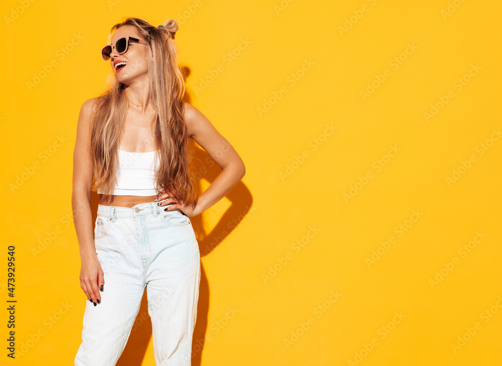 Portrait of young beautiful smiling blond female in trendy summer clothes. Sexy carefree woman posing near yellow wall in studio. Positive model having fun indoors. Cheerful and happy in sunglasses