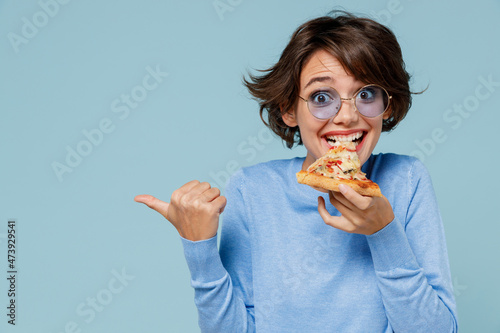 Young woman 20s in casual sweater biting eat slice of pizza point index finger aside on copy space area isolated on plain pastel light blue background studio portrait. People lifestyle food concept.