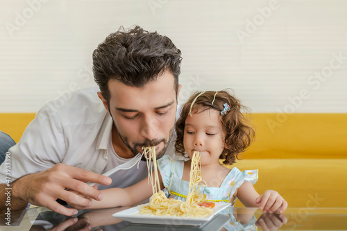 A cheerful young family eats spagetti with their daughter on a carpeted floor.