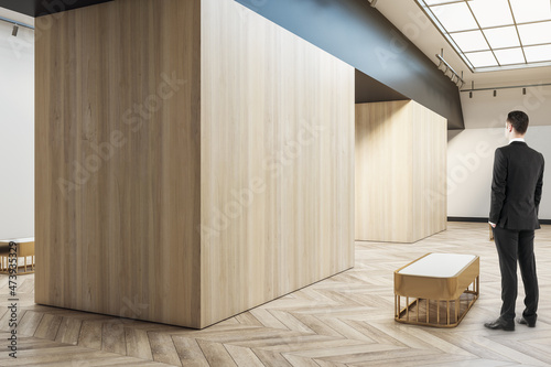 Man standing in modern wooden exhibition hall interior with sunlight from ceiling window, mock up place and seats. Gallery and museum concept.