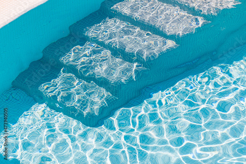 Closeup of swimming pool steps and clear blue water. New modern swimming pool entrance steps down with clean fresh refreshing blue water on bright hot summer day. Natural sunlight