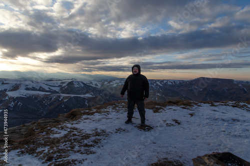 A man in warm winter clothes stands against the background of a mountain range in winter. The cloudy sky is colored by the rays of the setting evening sun