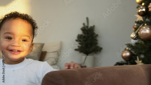 Early Christmas morning, a little boy runs around the sofa and hides behind it playing hide and seek. He smiles and has fun. The light of the sun falls on his face. There is a decorated Christmas tree