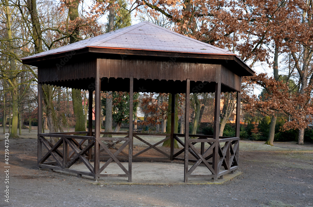 polygonal pergola with benches and a fence around the perimeter. there is a lot of space inside. the roof is pyramidal covered with asphalt shingles, in natural park, footpath, gravel