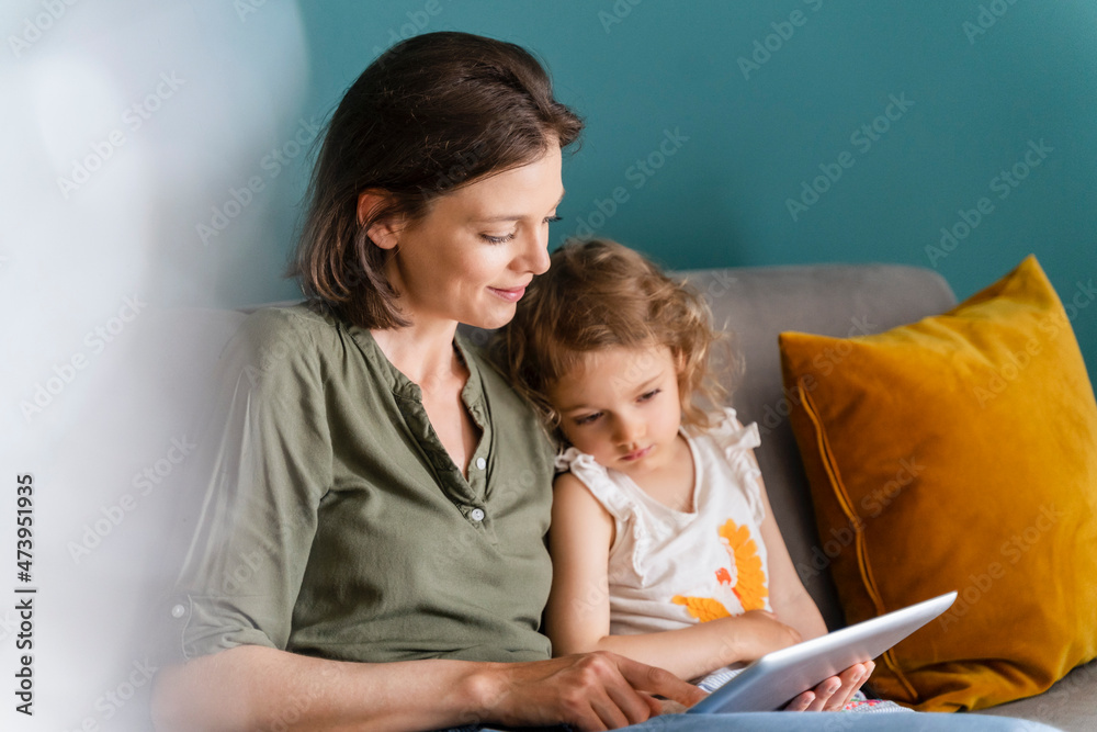 Mother using digital tablet while sitting with daughter at home Mother using digital tablet while sitting with daughter at home