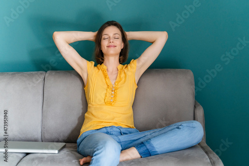 Woman with hands behind head sitting by laptop on sofa at home photo