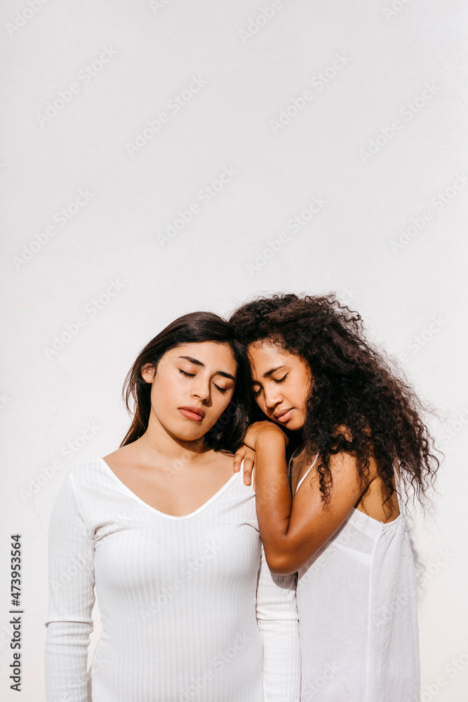 Female friends with eyes closed leaning on each other in front of white wall Female friends with eyes closed leaning on each other in front of white wall