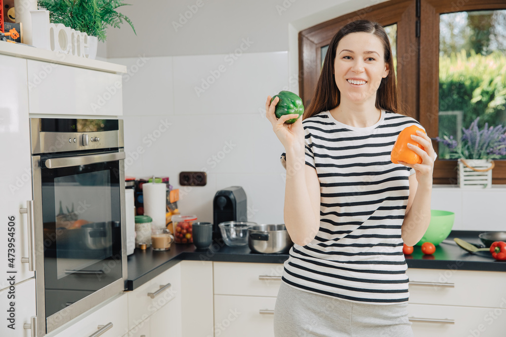 Healthy lifestyle. Woman stands in the kitchen and holds two peppers in her hands. Green and orange peppers for cooking. Cooking with natural ingredients Healthy lifestyle. Woman stands in the kitchen and holds two peppers in her hands. Green and orange peppers for cooking. Cooking with natural ingredients