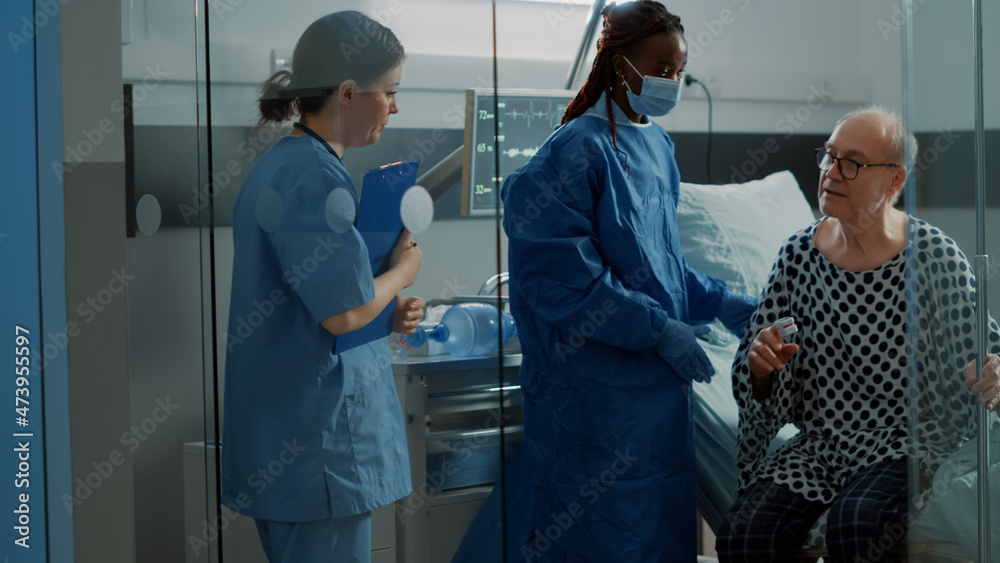 Obraz premium African american surgeon walking elder patient from hospital ward with nurse at intensive care facility. Sick old man connected to modern oximeter and IV drip bag leaving bed for surgery