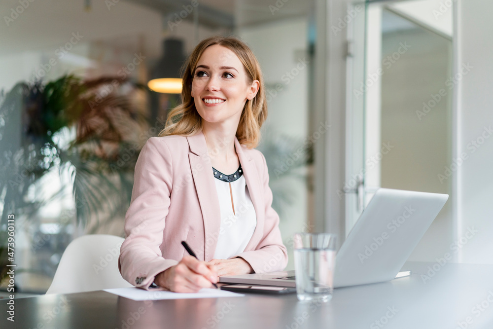 Smiling female professional looking away while writing on document in front of laptop at desk Smiling female professional looking away while writing on document in front of laptop at desk