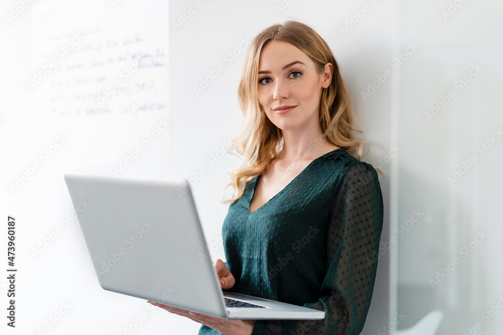Female professional with laptop leaning on wall in office Female professional with laptop leaning on wall in office