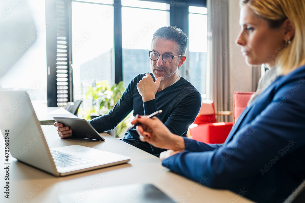 Mature businessman looking at laptop while discussing with female coworker in creative office Mature businessman looking at laptop while discussing with female coworker in creative office