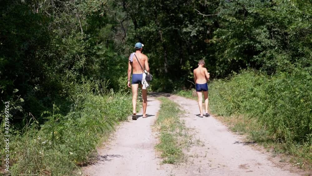 Dad and Son are Walking Along a Sandy Path in a Green Woods. Back view of naked man and child in swimming trunks taking sunbaths, walking outdoors. Summertime. Relaxation. Happy family. Rest. Relax. Dad and Son are Walking Along a Sandy Path in a Green Woods. Back view of naked man and child in swimming trunks taking sunbaths, walking outdoors. Summertime. Relaxation. Happy family. Rest. Relax.