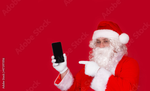 Portrait of a cheerful Santa Claus showing a device, a gadget screen, a smartphone, advertising services and goods, isolated on a bright red background