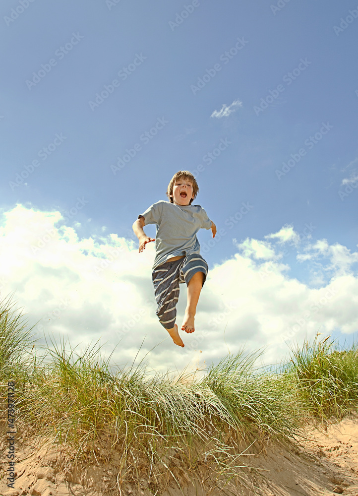Carefree boy jumping over sand dunes at beach during sunny day Carefree boy jumping over sand dunes at beach during sunny day