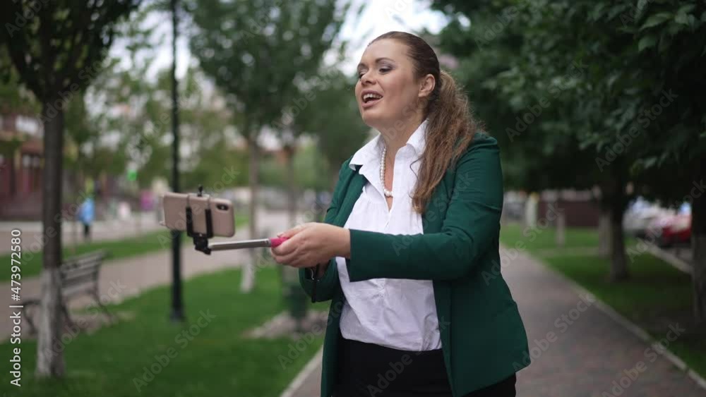 Positive overweight female business blogger standing in city talking smiling waving greeting subscribers in social media on smartphone. Happy confident beautiful Caucasian woman blogging outdoors
