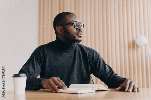 Thoughtful african businessman sits at desk with pen and agenda looks away with pensive expression