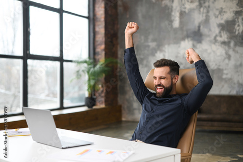 Excited young businessman in smart casual wear celebrating successful deal, looking at monitor raising fists in triumph, overjoyed male entrepreneur done project, screaming yes sitting at office photo