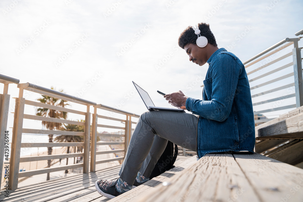 Afro man using smart phone over laptop while sitting on steps at promenade Afro man using smart phone over laptop while sitting on steps at promenade