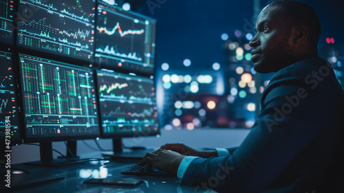 Financial Analyst Working on Computer with Multi-Monitor Workstation with Real-Time Stocks, Commodities and Exchange Market Charts. African American Businessman Works in Investment Bank Late at Night.