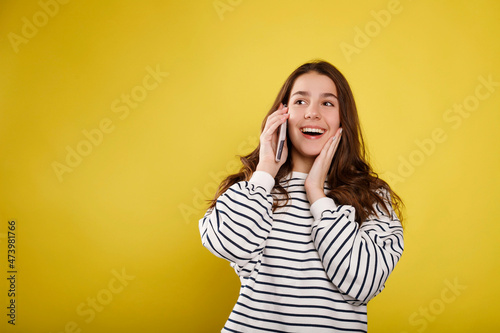Happy young teenager girl talking on the phone, cheerful woman enjoys pleasant mobile conversation, smiling millennial female holding cell speaking making call by telephone over yellow background