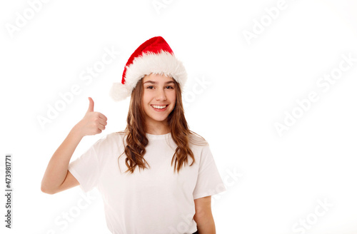 Young happy teenager girl in Santa Claus red hat standing over white background smiling with happy face showing hand ok sign. Christmas and New Year lifestyle concept