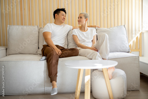 Multiracial couple sit and rest on sofa at home. Concept of relationship and spend time together. Idea of domestic lifestyle. Asian man and caucasian woman looking in sunny window in studio apartment