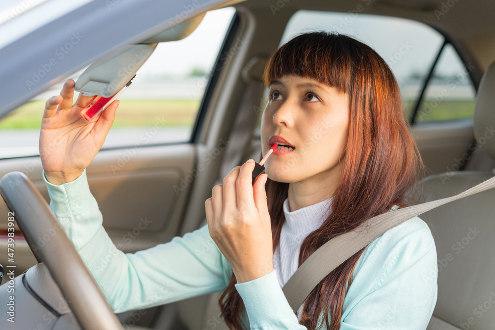 Young woman looking in rear view mirror and making up her lips while sitting behind the wheel of her car. Female painting lips doing applying make up while driving the car. Concept of danger driving.